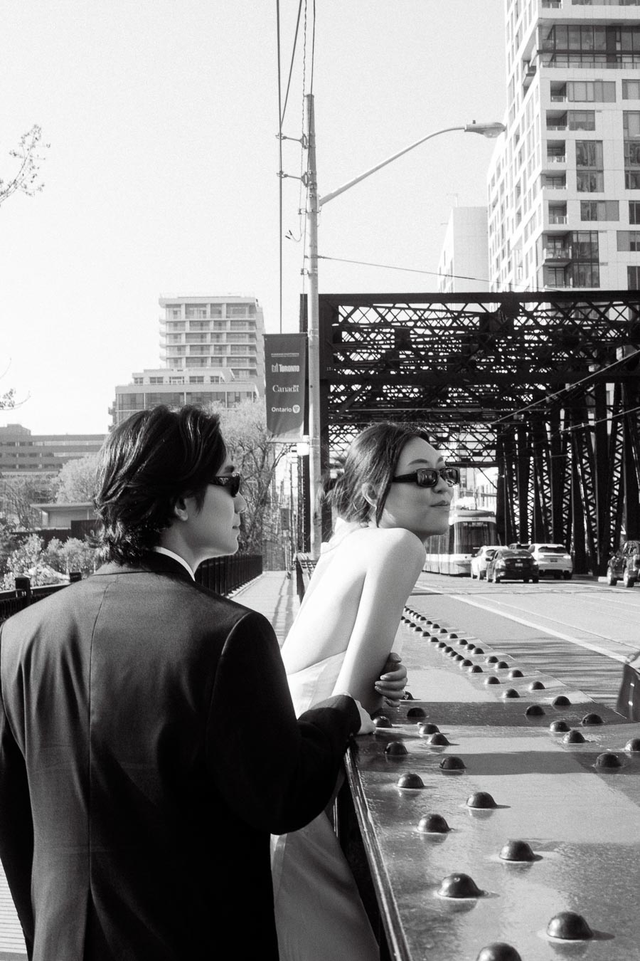 A couple in formal clothing and sunglasses stand together on a city bridge, with modern buildings and streetcars in the background.