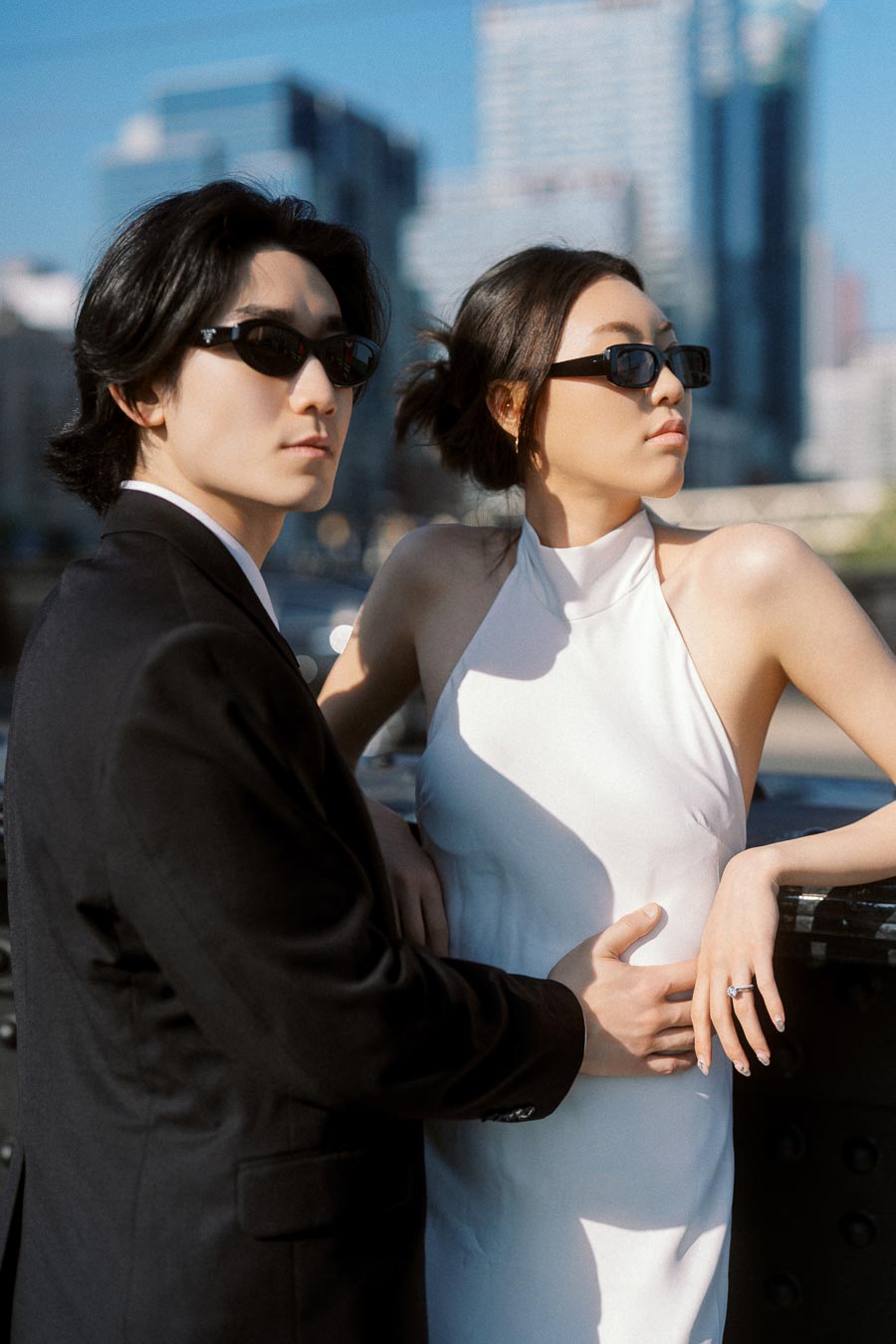 A fashionable couple in black suit and white dress, both wearing sunglasses, embrace on a downtown Toronto bridge with city skyscrapers behind them.