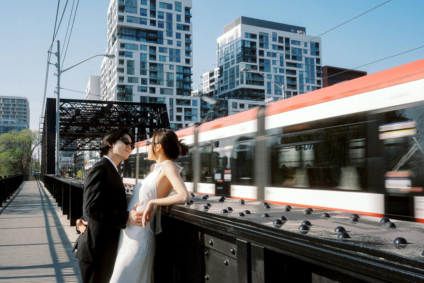 Couple in formal attire leaning on Sir Isaac Brock Bridge, with a moving streetcar and modern city buildings in the background.