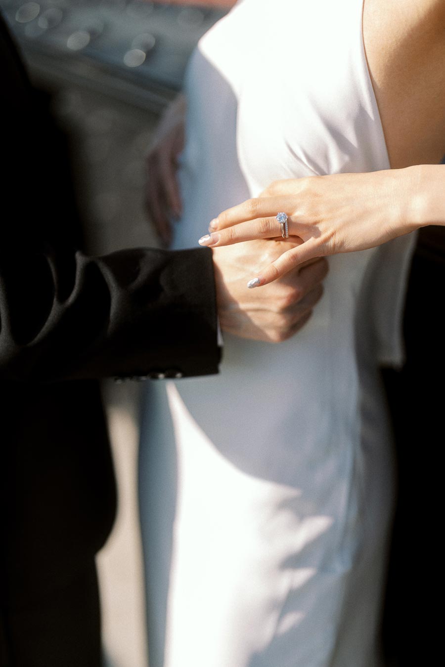 Close-up of a woman's hand with an engagement ring resting on her partner's arm, both dressed formally.