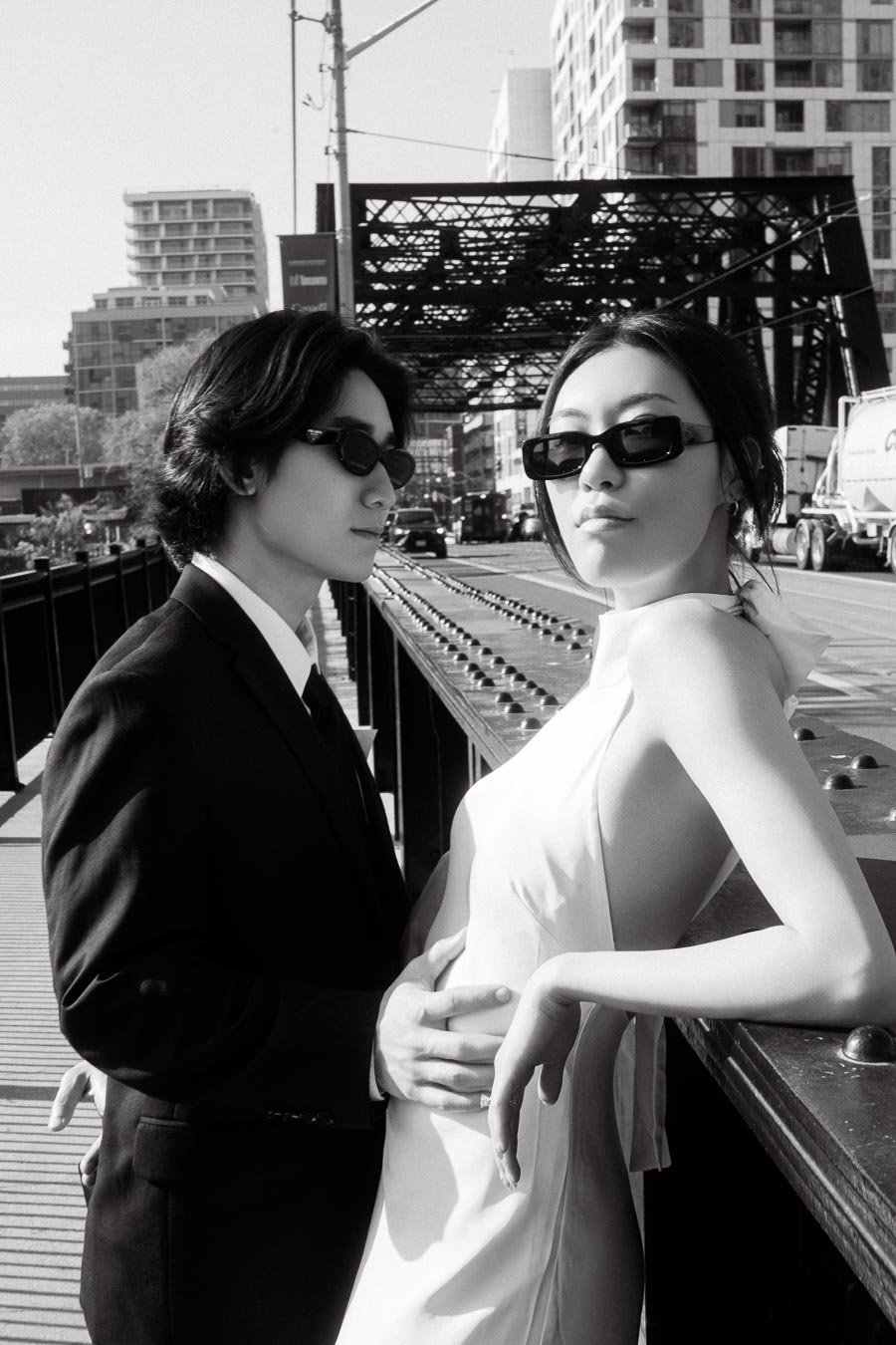 Couple wearing sunglasses, one in a suit and one in a white dress, posing closely on an industrial bridge in downtown Toronto.