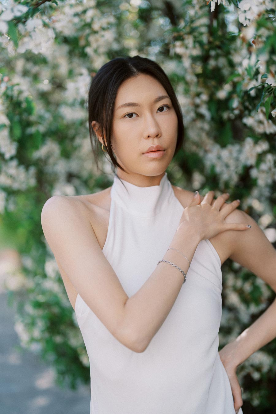 Woman in a sleeveless white dress stands in front of flowering branches, posing serenely with one hand on her shoulder.