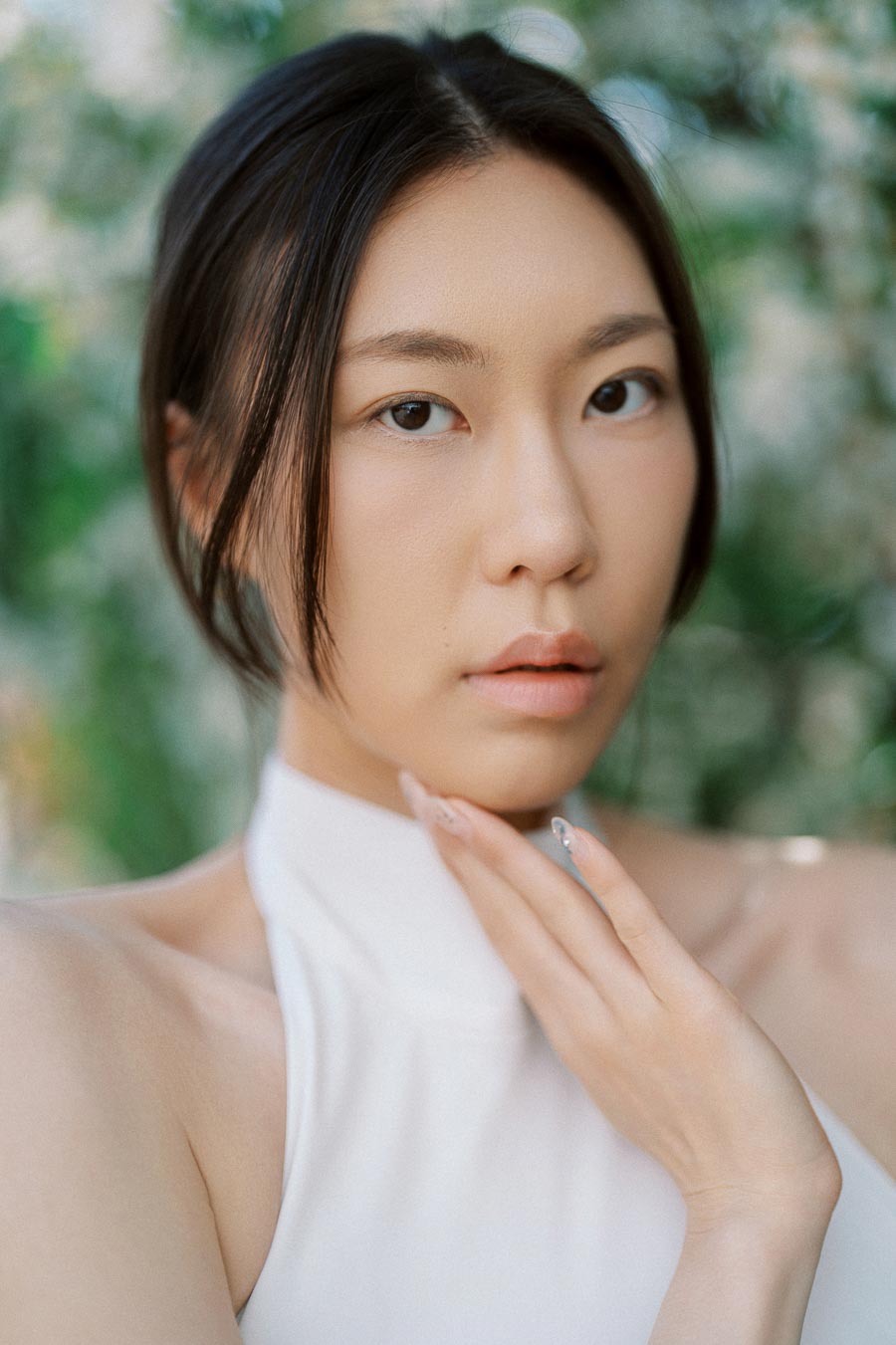 Woman in white halter dress gazing softly at the camera, hand gently touching chin, with blurred greenery behind her.
