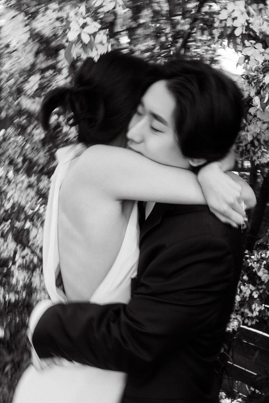 Couple embracing closely in front of blooming branches, one in a white dress and the other in a black suit, with soft focus and romantic mood.
