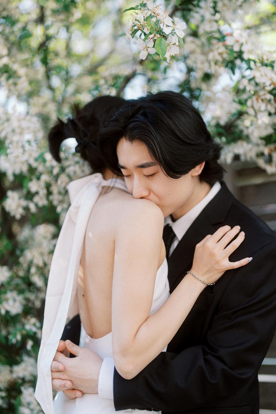 Couple in formal attire embracing closely, one kissing the other's shoulder, surrounded by white spring blossoms outdoors.