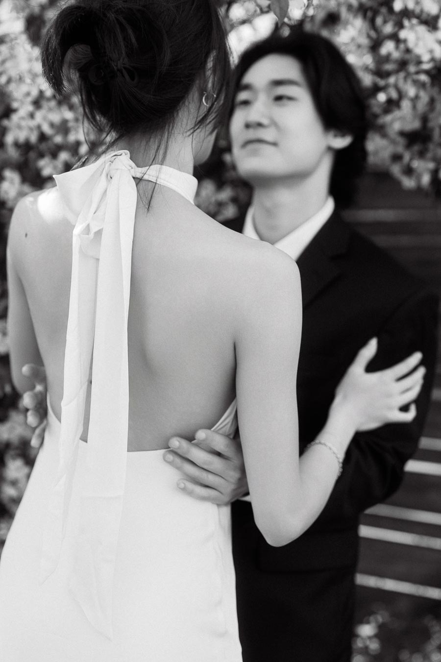 Couple in formal attire embracing closely under blooming branches, with the woman's back to the camera and the man facing her.
