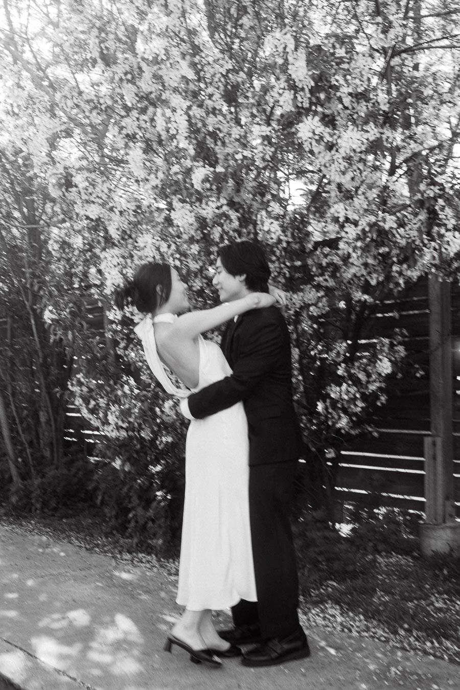 Couple in formal attire embracing closely on a sidewalk with flowering trees in the background.