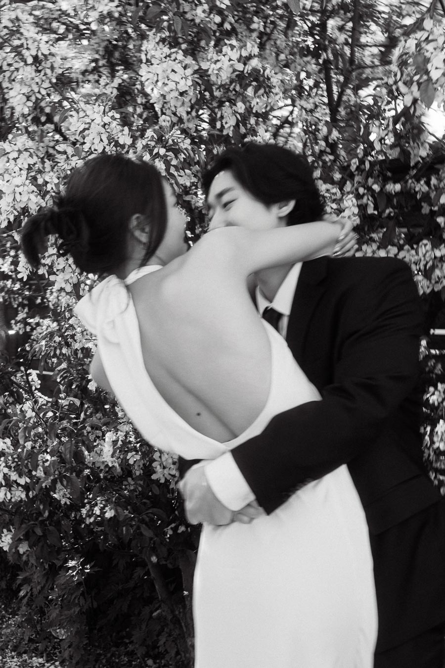 Couple in a loving embrace, one in a white dress and the other in a dark suit, surrounded by blooming bushes outdoors.