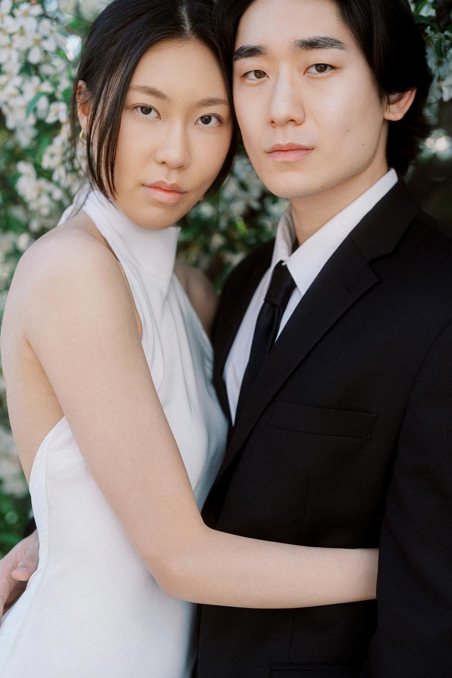Couple embracing in formal attire, surrounded by white blossoms, gazing softly at the camera