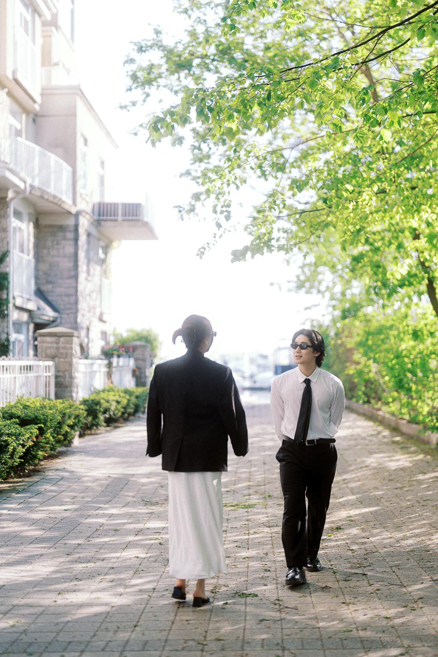 Two people in formal attire walking toward each other on a leafy, sunlit city path with greenery and buildings around them.