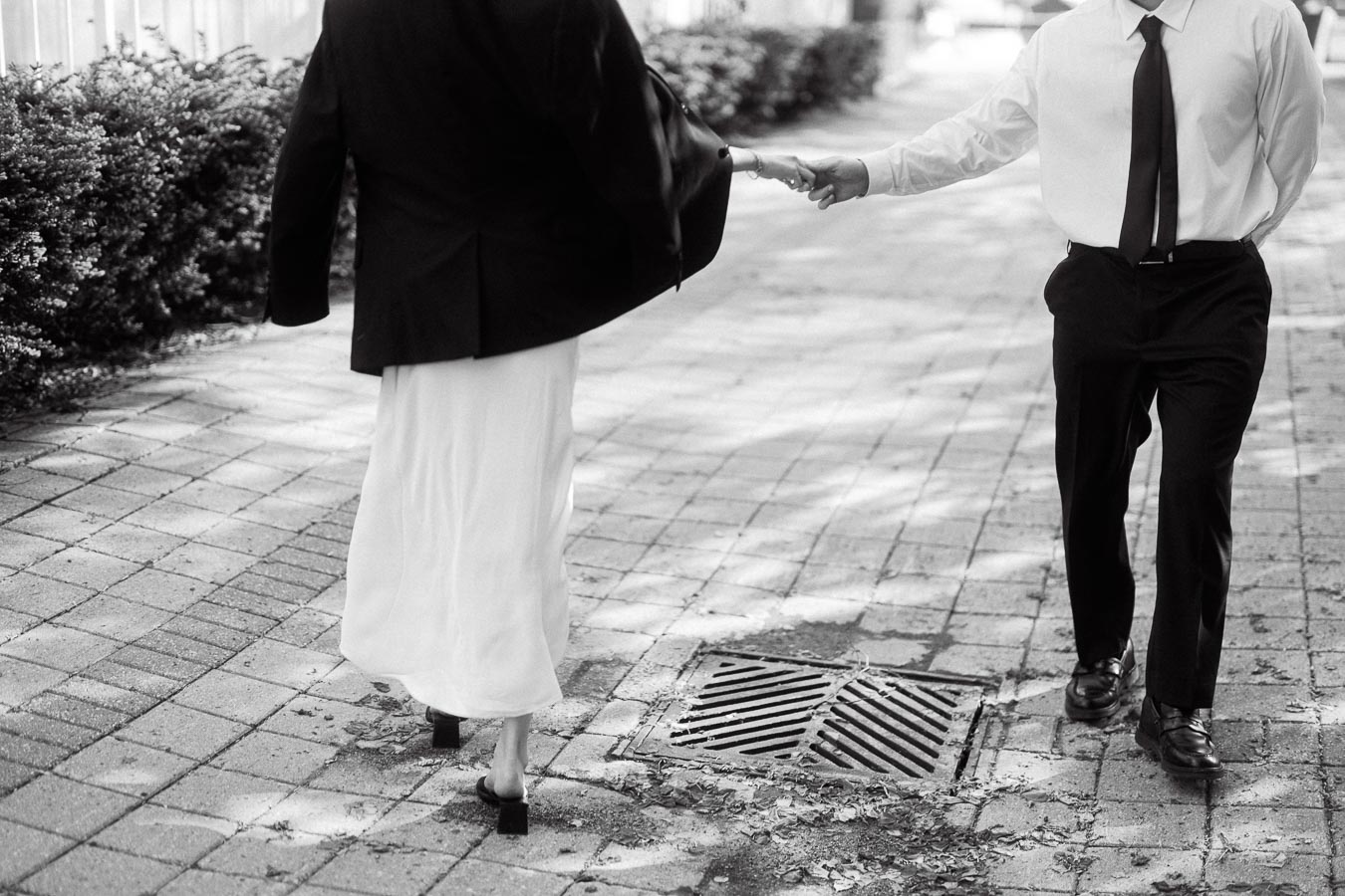 Couple holding hands while walking on a paved path, dressed in black and white, sunlight filtering through trees.