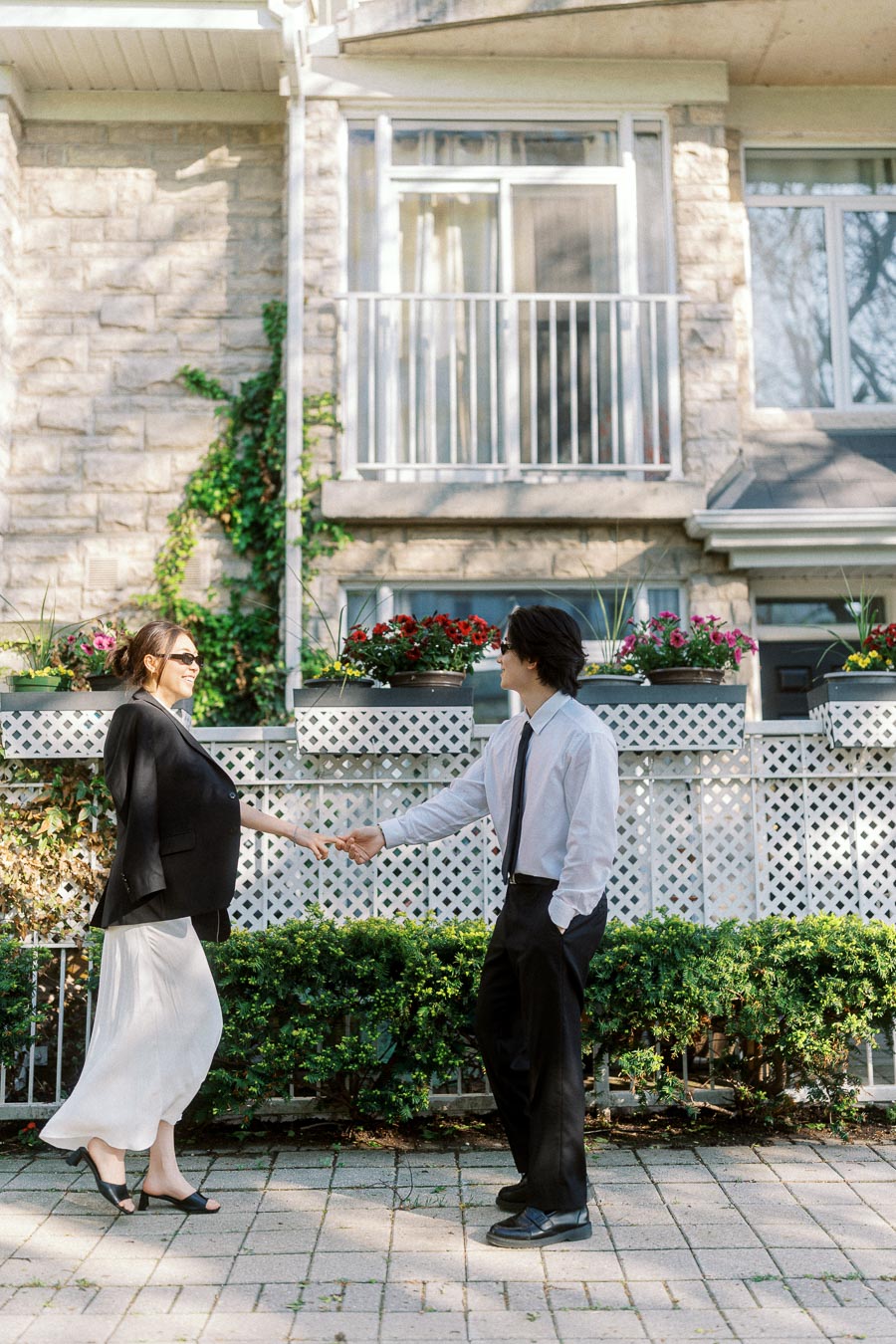 Couple holding hands and smiling in front of a stone townhouse with flower boxes and greenery.