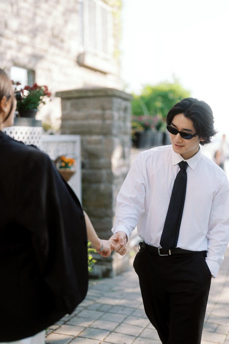 Person in white shirt and black tie wearing sunglasses holds hands with partner while walking outdoors on a bright day.
