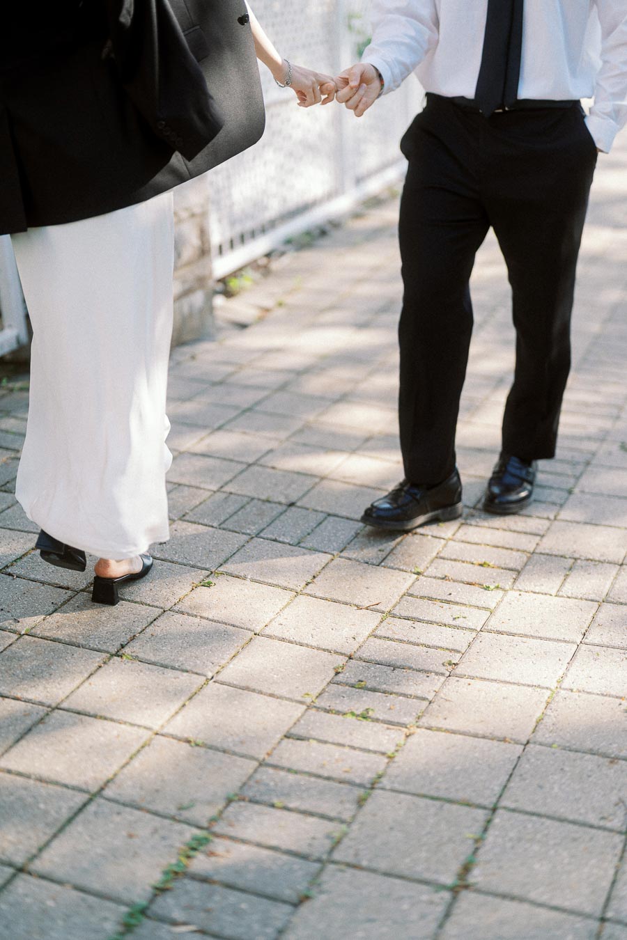 Couple holding hands while walking on a paved sidewalk, dressed in black and white formal outfits, sunlight casting soft shadows.