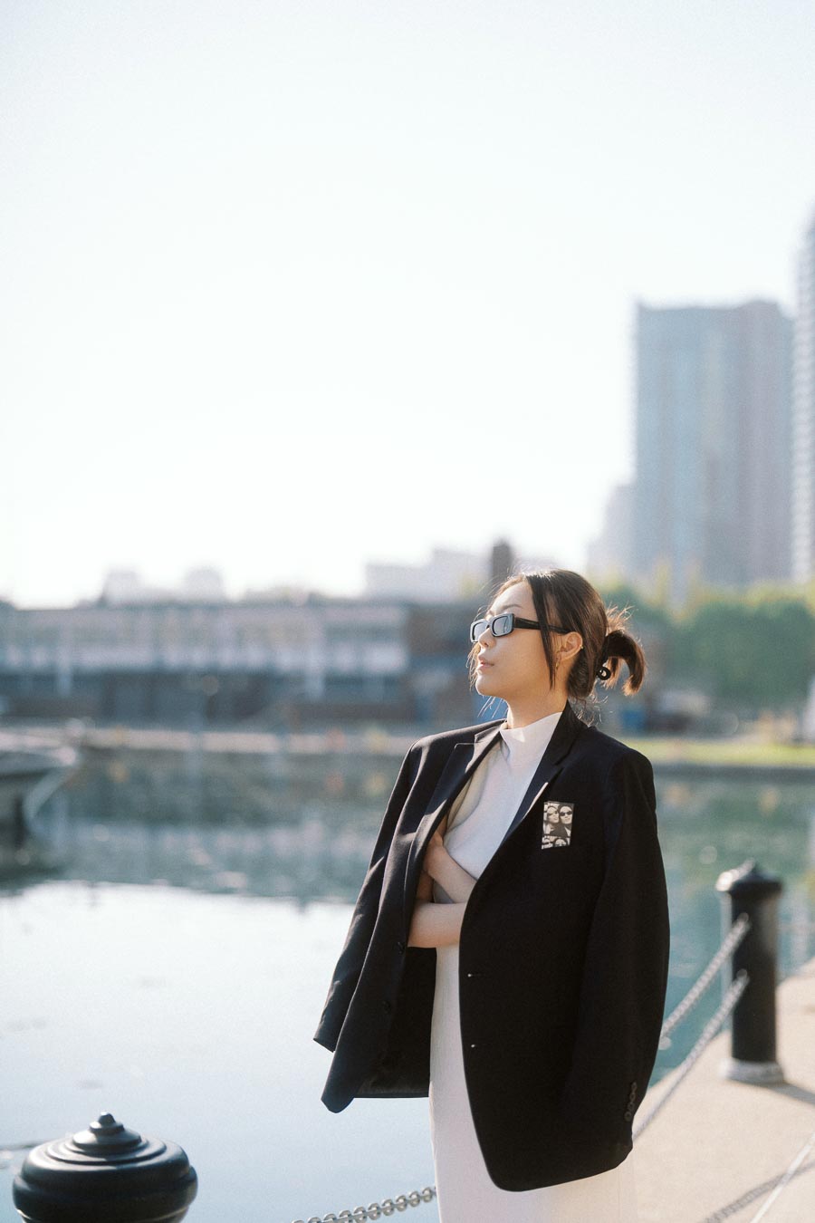 Woman wearing sunglasses and a black blazer over a white dress standing near a city waterfront with soft light and buildings in the background.