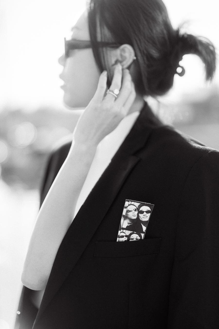 Close-up of a person in a black suit and sunglasses, touching their ear, with a photo booth strip in the pocket.