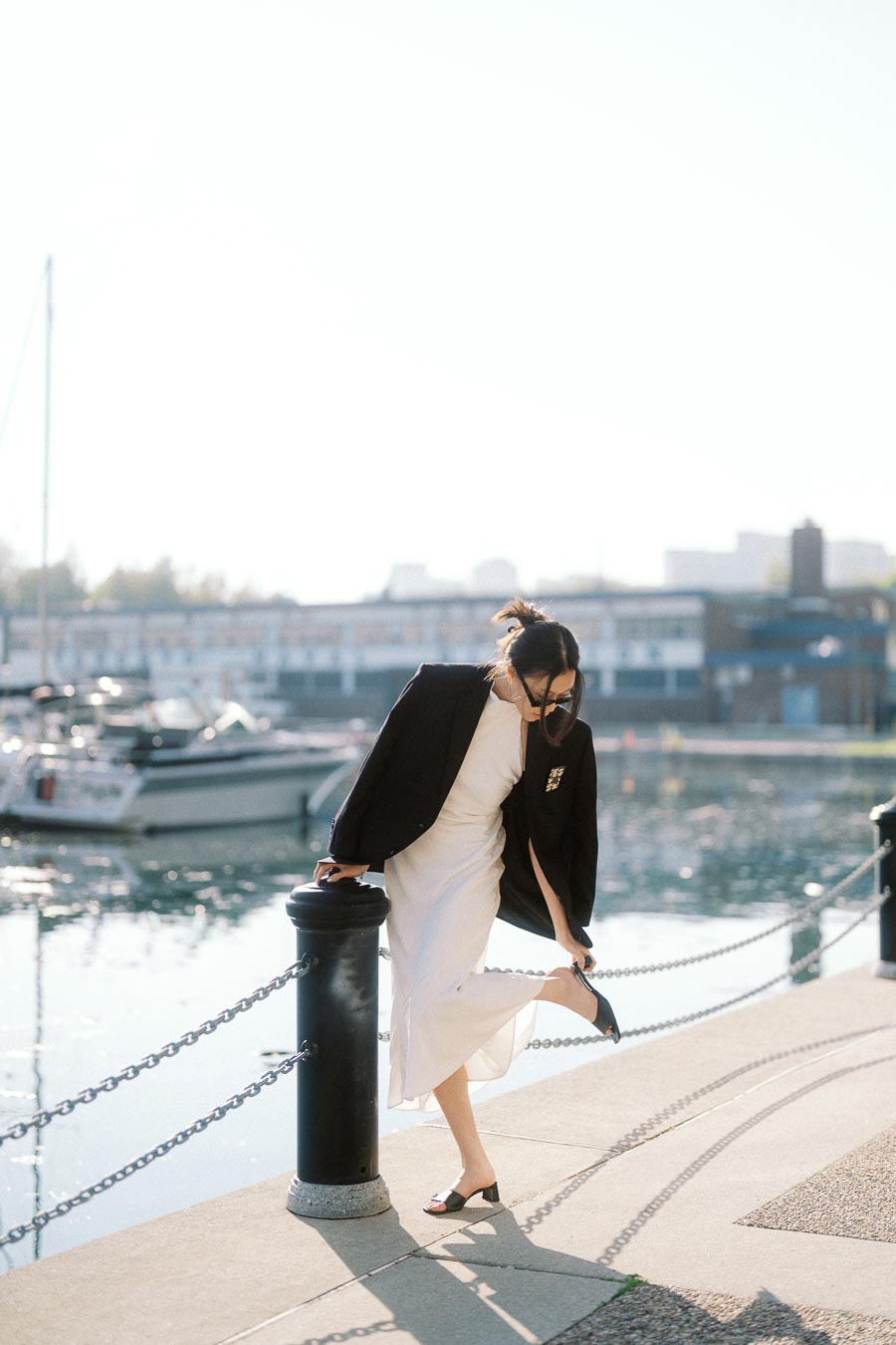 Woman in white dress and black blazer adjusting her shoe beside a marina with boats and sparkling water.