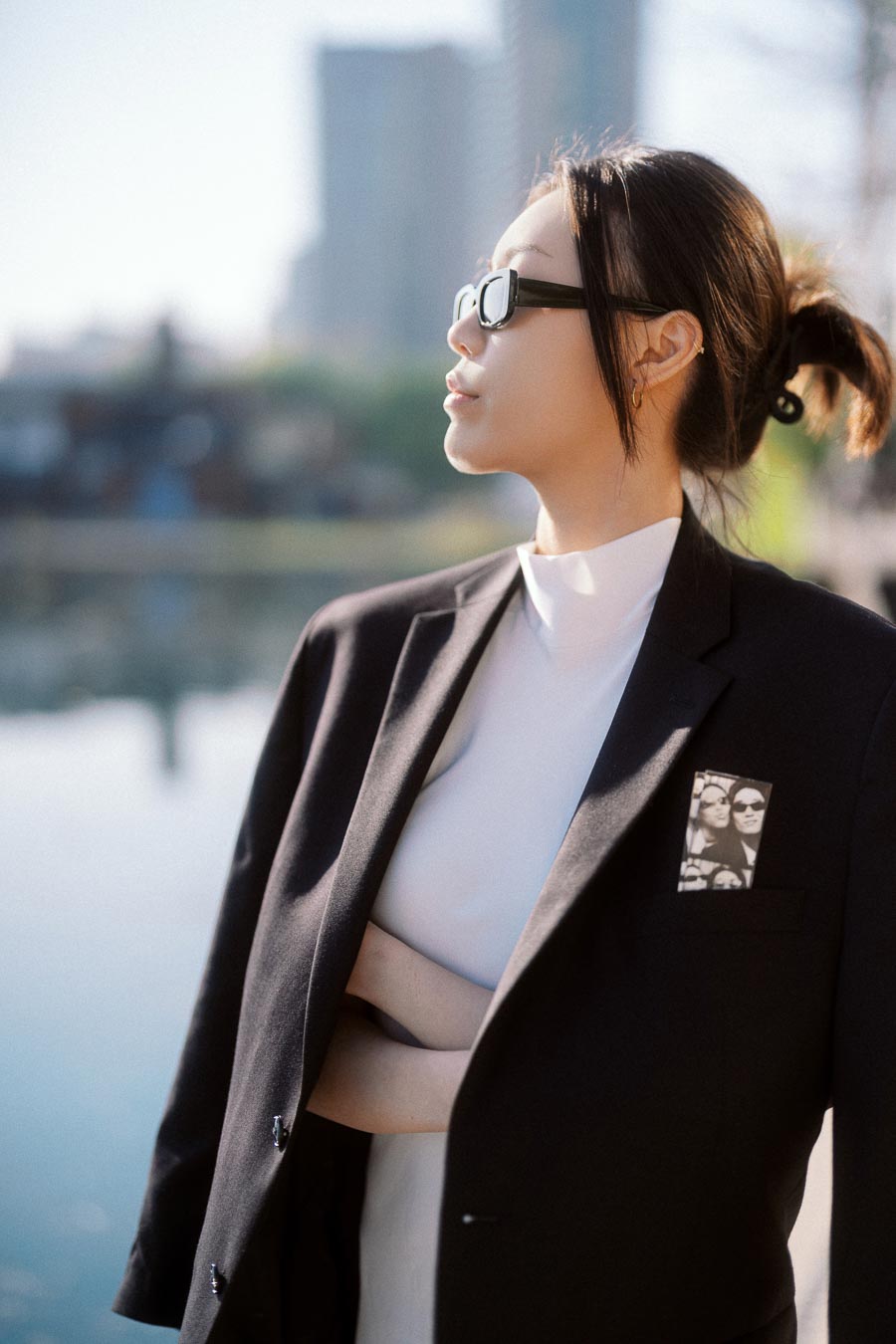 Person in black blazer and sunglasses stands by waterfront with blurred city skyline behind them.