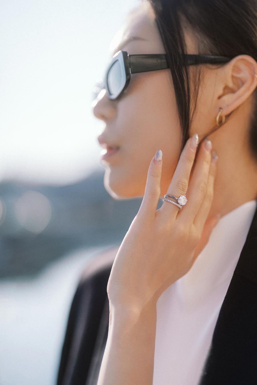 Close-up of a woman in sunglasses showing her engagement ring near her face, with soft sunlight and blurred water background.