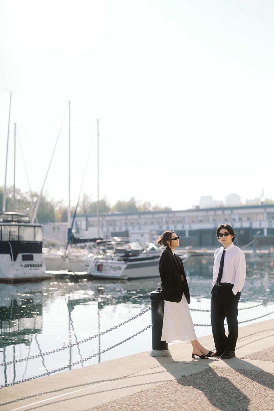 Two people in formal attire and sunglasses standing near a marina with boats, sunlight reflecting on the water.
