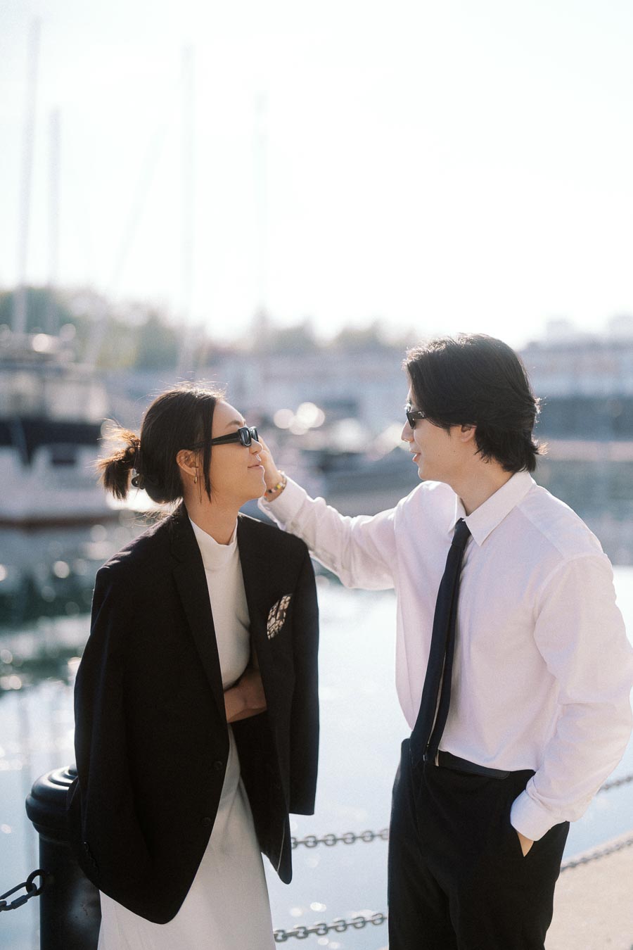 Couple in stylish black and white outfits smiling at each other by a marina with boats in the background.