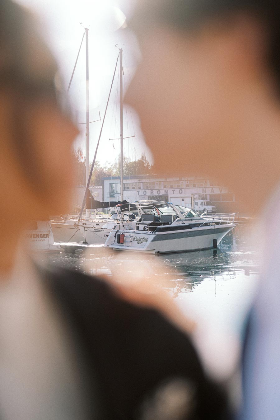 Sailboats docked at a marina with a couple's blurred silhouettes in the foreground, sunlight reflecting on the water.