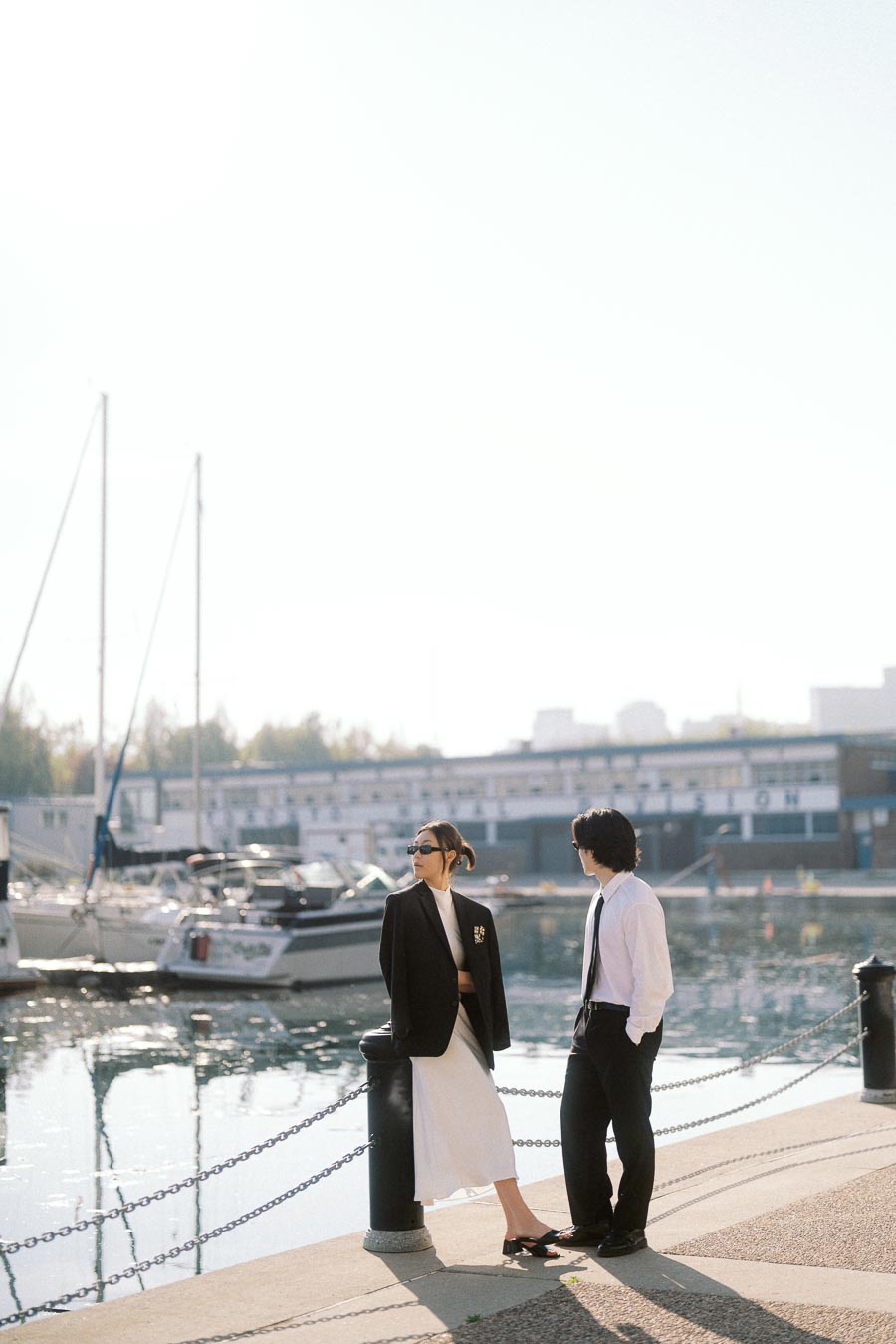 A couple in formal attire stands near the marina waterfront with boats and calm water behind them on a sunny day.