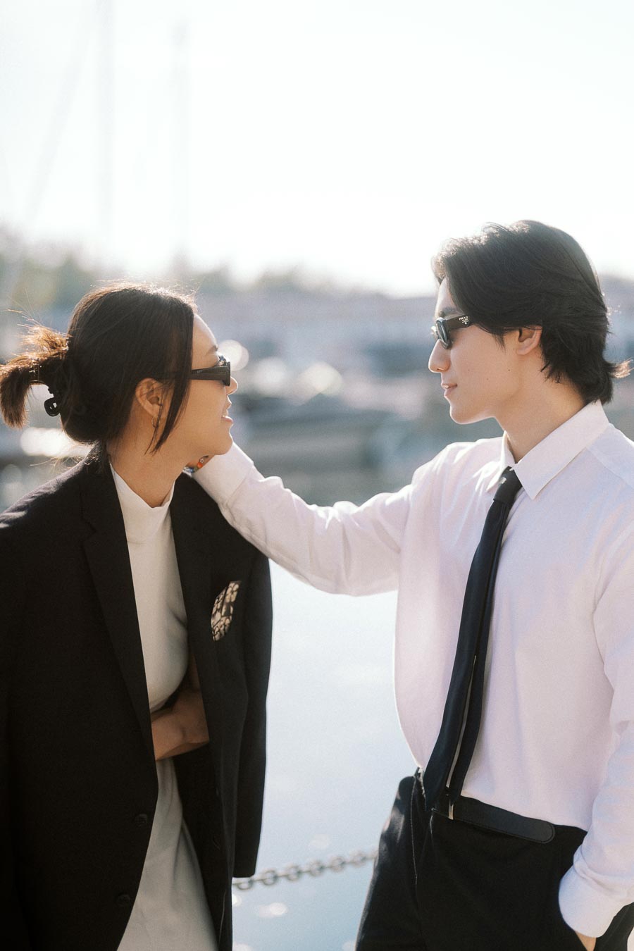 Couple wearing sunglasses and formal outfits looking at each other affectionately by the marina waterfront.