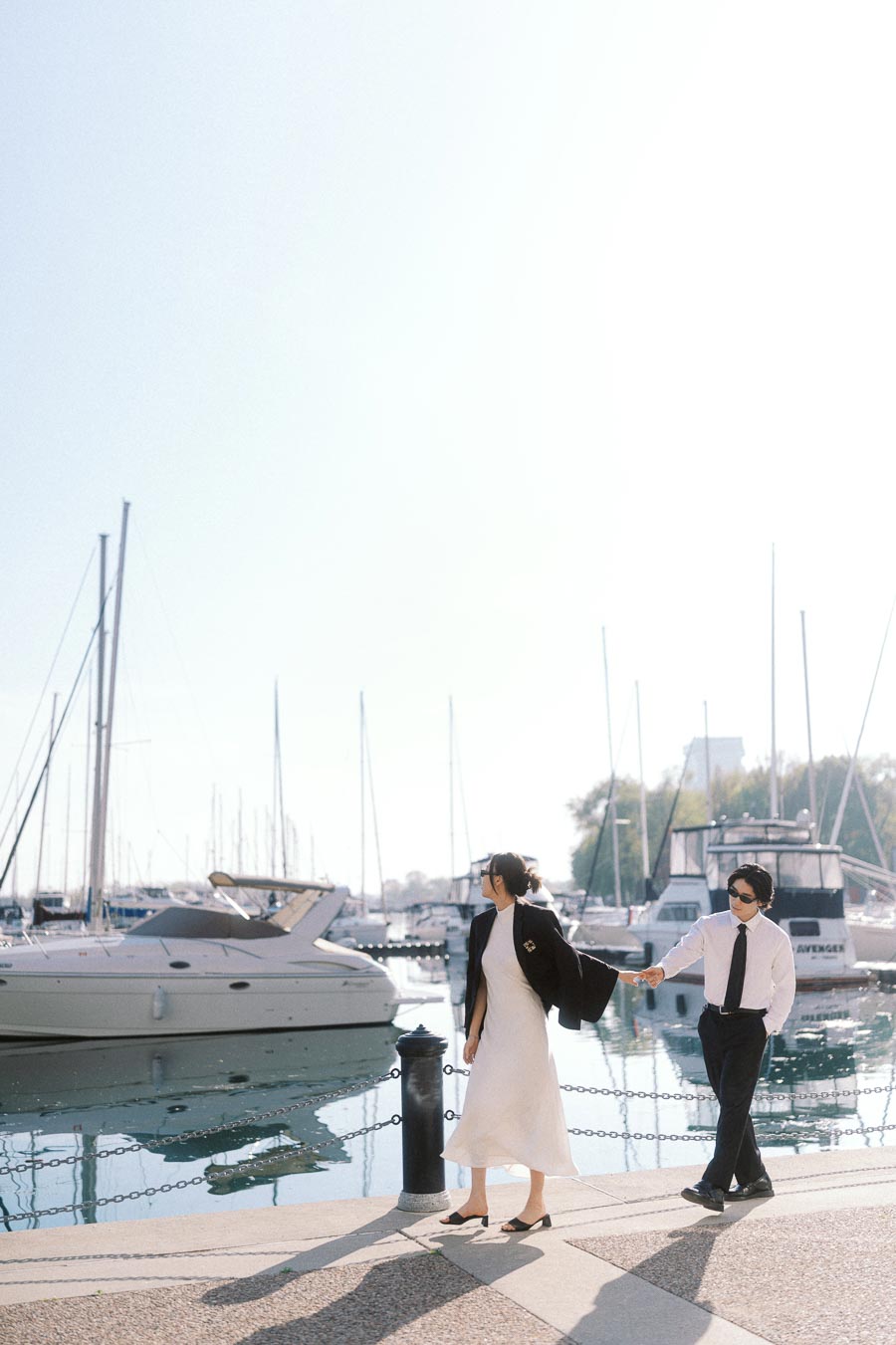 Couple dressed in black and white holding hands by a marina with boats and calm water on a sunny day.