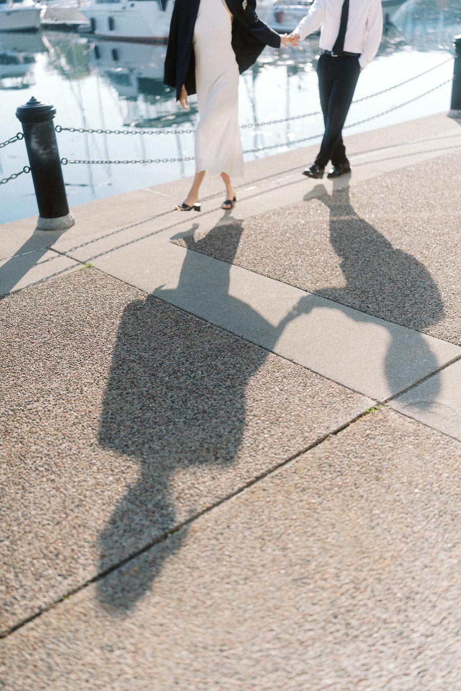 Shadows of a couple holding hands while walking along a marina waterfront with boats reflected in the water.