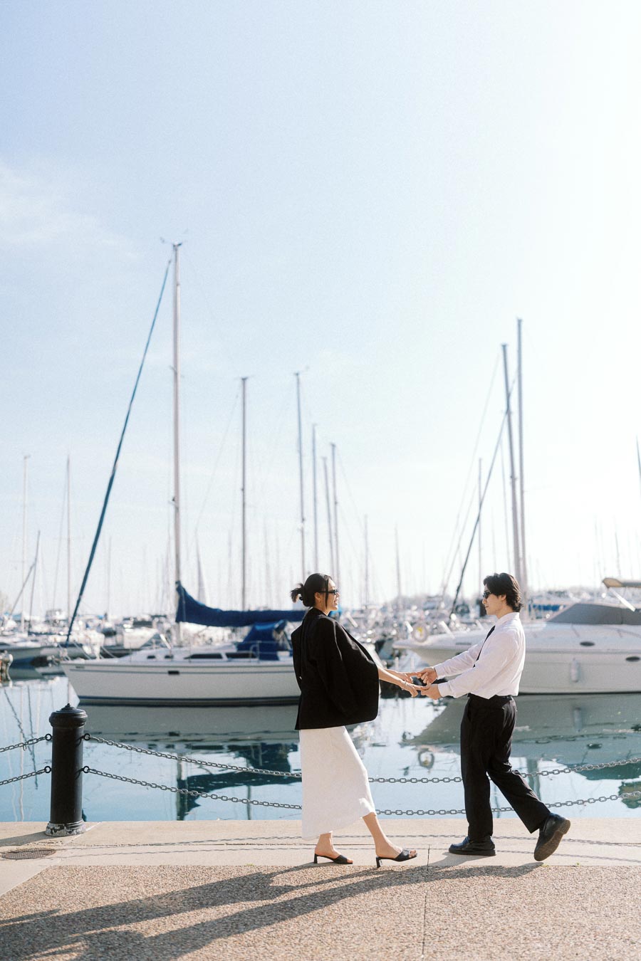 Couple holding hands and smiling by a marina with sailboats in the background on a sunny day.