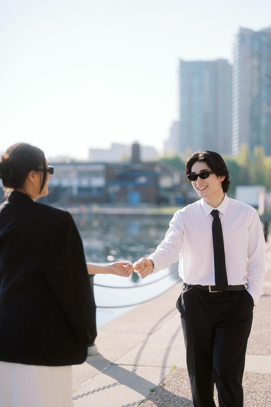 Couple holding hands and smiling while walking along the waterfront in downtown Toronto, both wearing sunglasses and stylish black and white outfits.