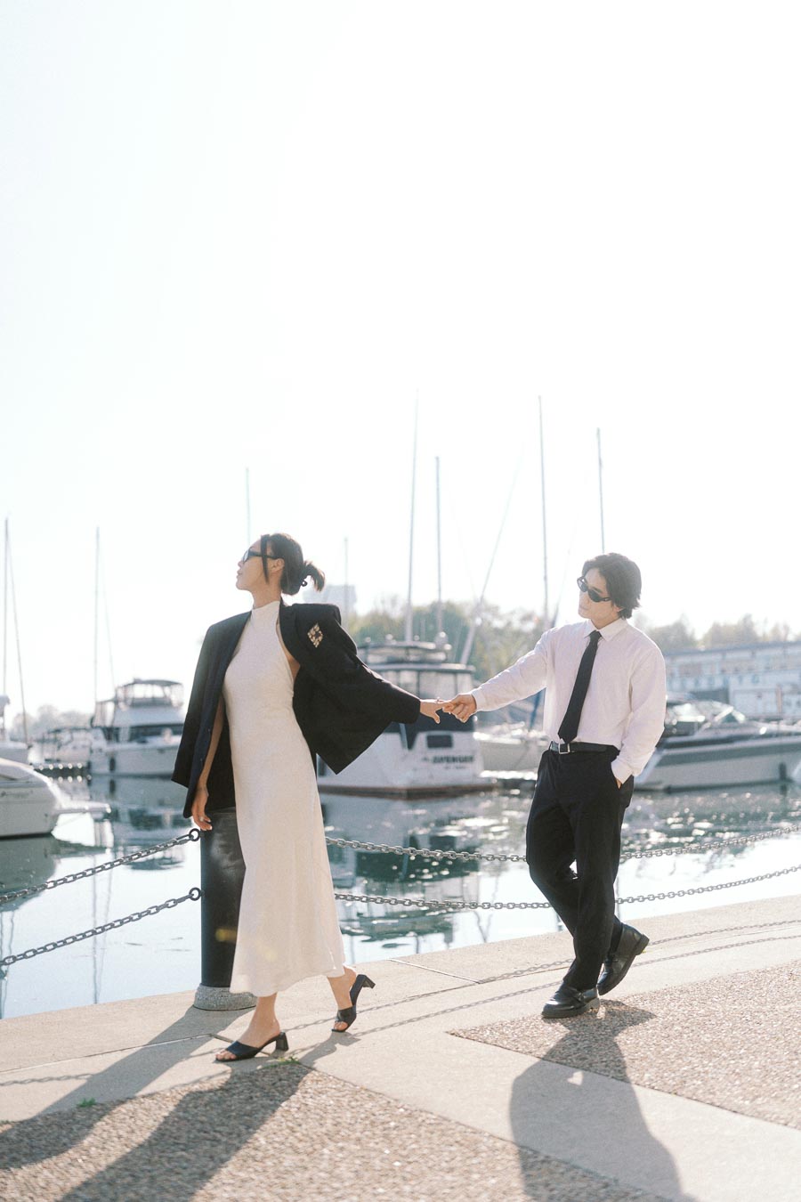 Couple in formal outfits holding hands while walking along a marina waterfront with boats and sunlight in the background.