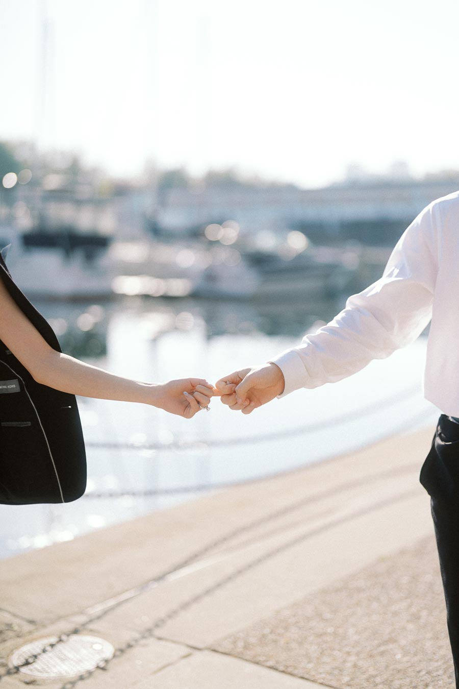 Close-up of a couple holding hands beside a marina with boats and sunlight reflecting on the water.