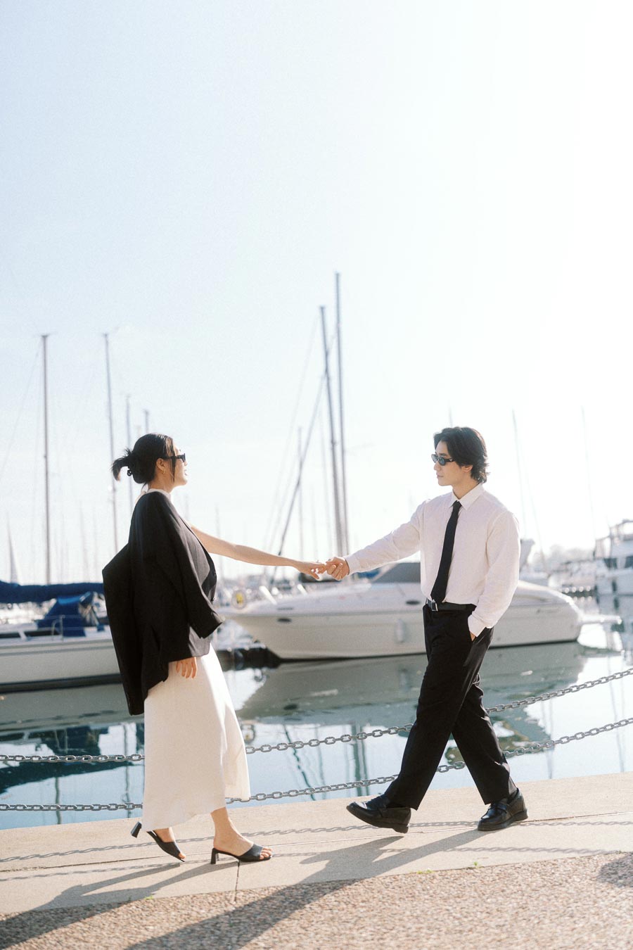 Couple holding hands and walking by the marina, dressed stylishly in black and white, with sailboats in the background.