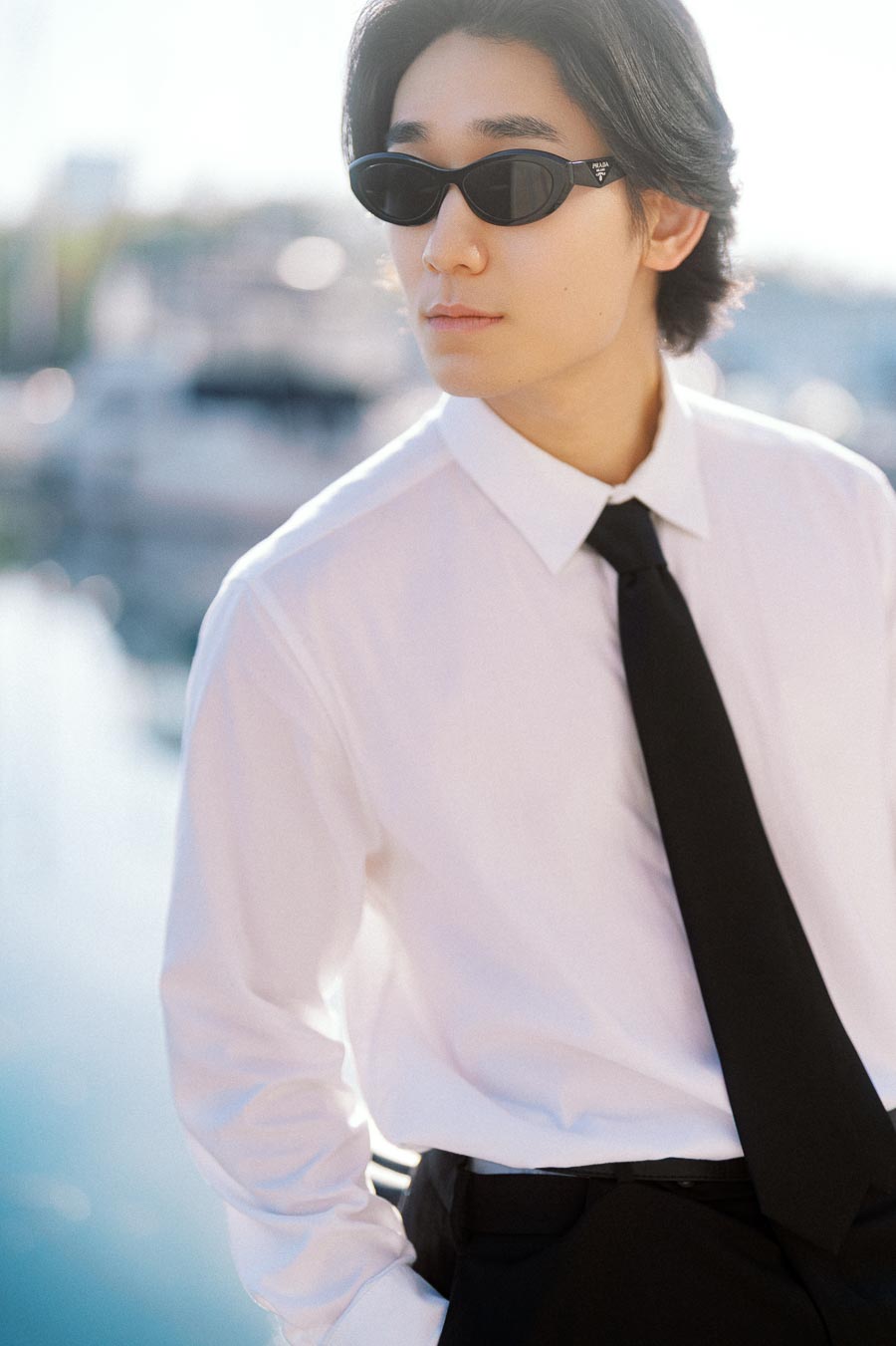 Person in white shirt, black tie, and sunglasses standing by a marina with boats in the background.