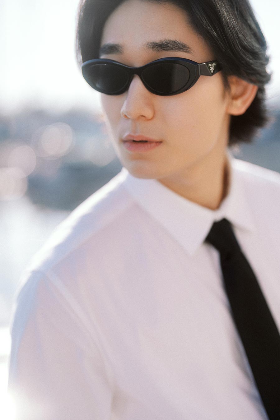 Person wearing black sunglasses, white shirt, and black tie looking away in soft outdoor light, urban background blurred.