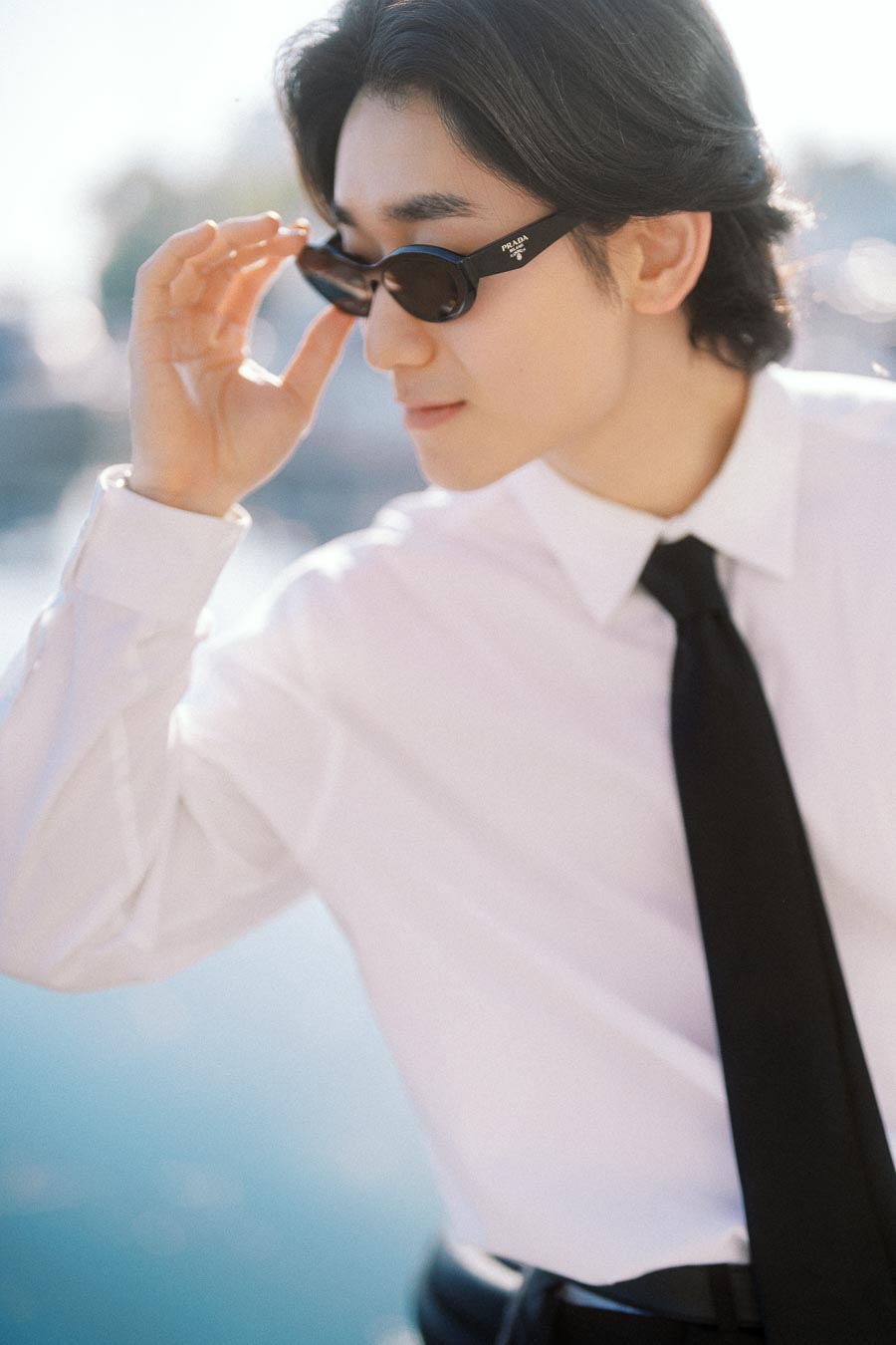 Person wearing sunglasses, white shirt, and black tie, adjusting their glasses with a blurred waterfront background.