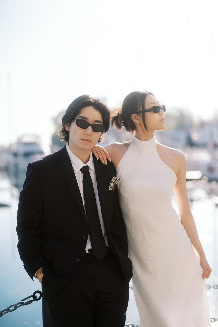 Couple wearing sunglasses, one in a black suit and the other in a white dress, standing together by the marina with boats in the background.