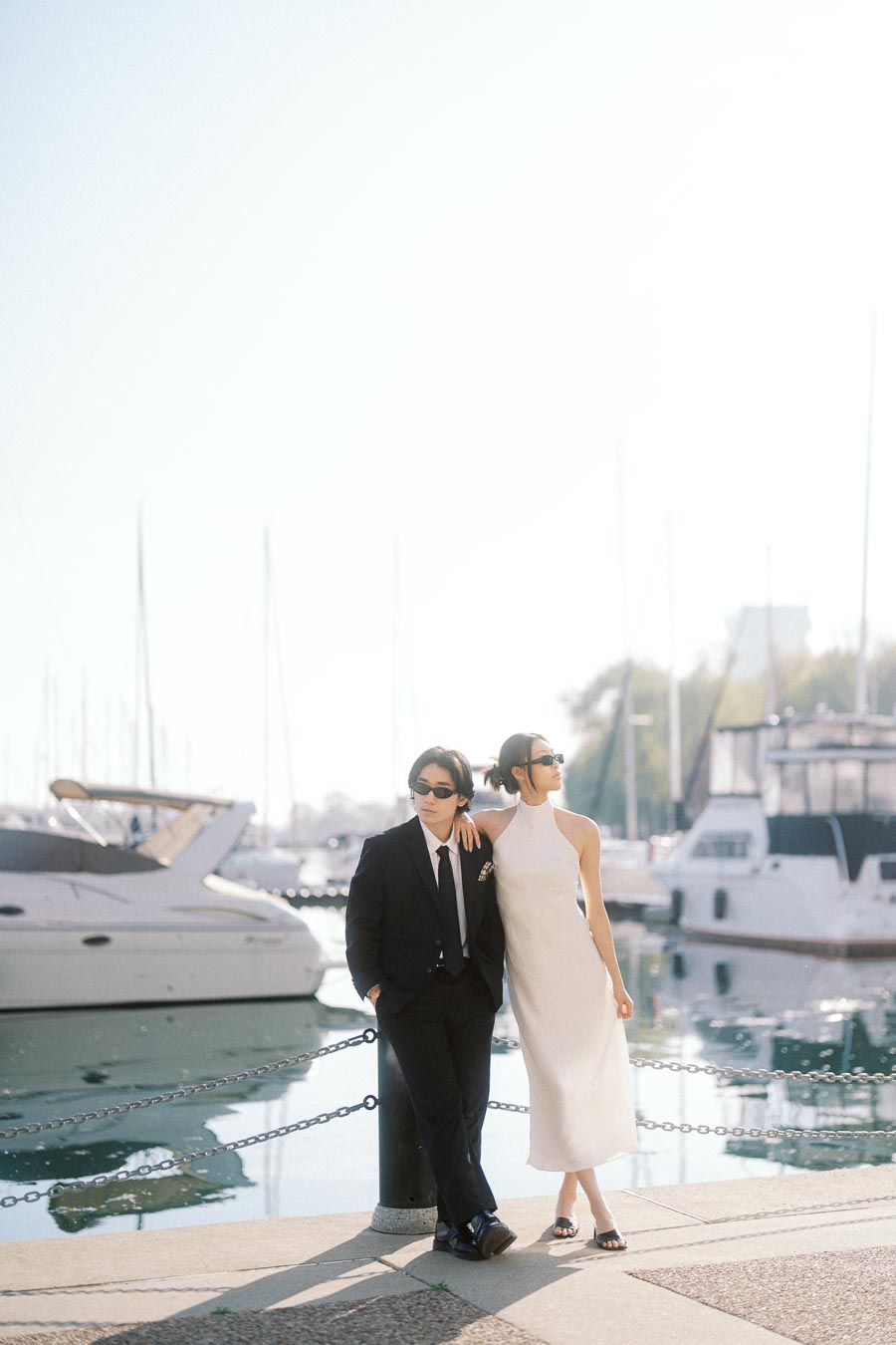 Couple in formal outfits and sunglasses standing by marina with yachts and calm water in Toronto.