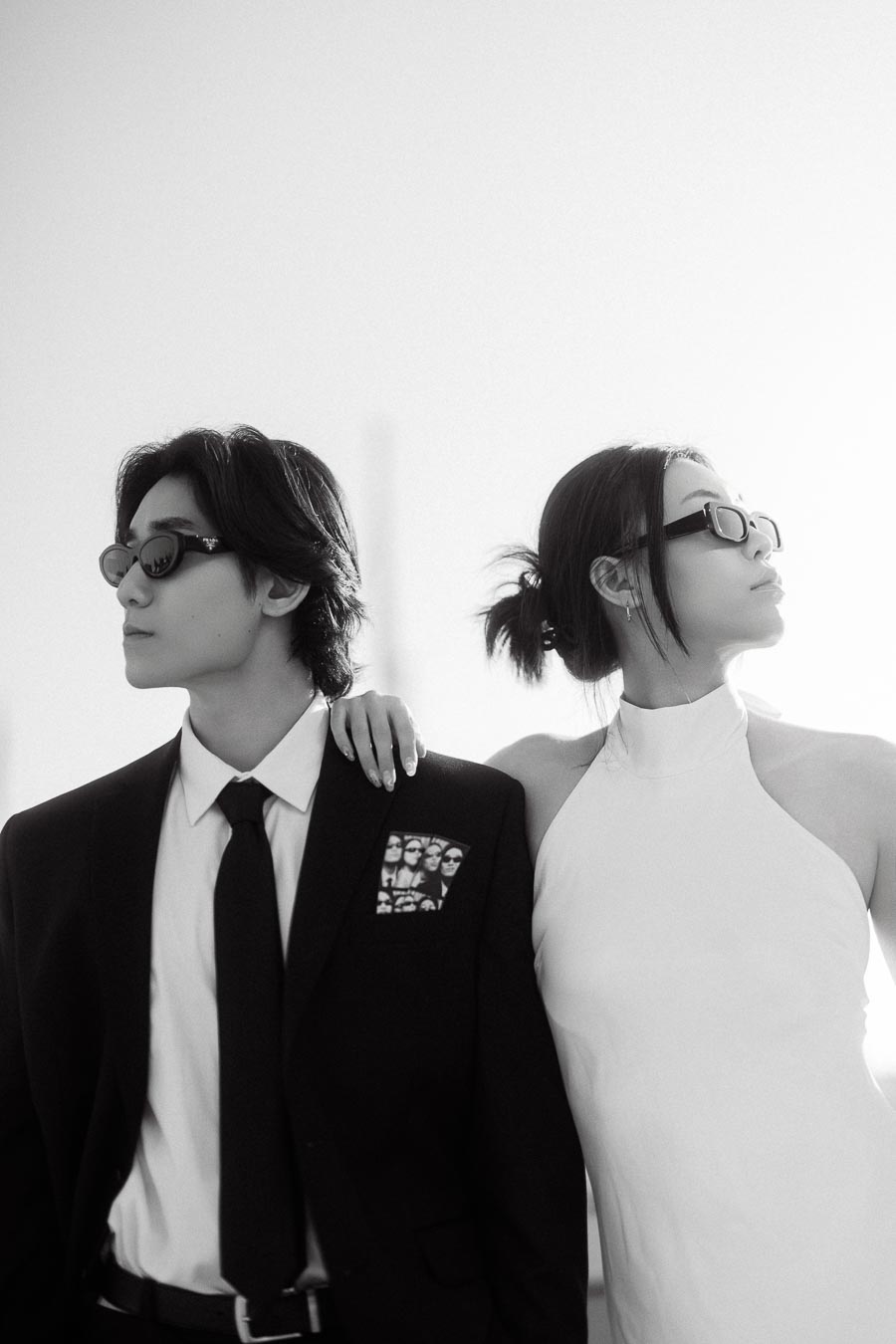 Couple wearing sunglasses, one in a black suit and the other in a white dress, posing stylishly outdoors in bright city light.