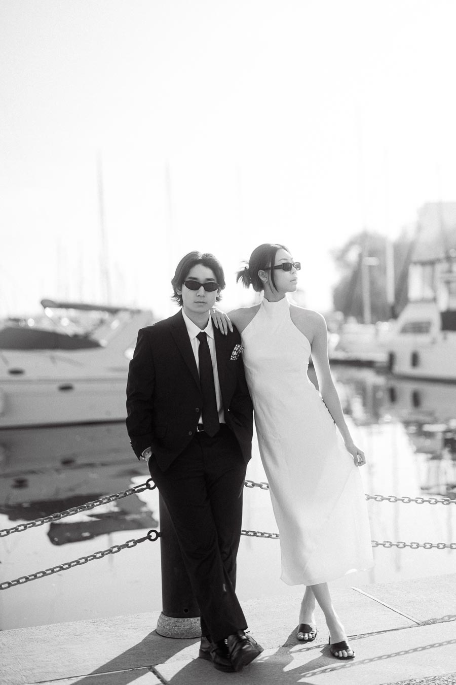 Couple in formal attire and sunglasses standing together by marina boats on a sunny day.