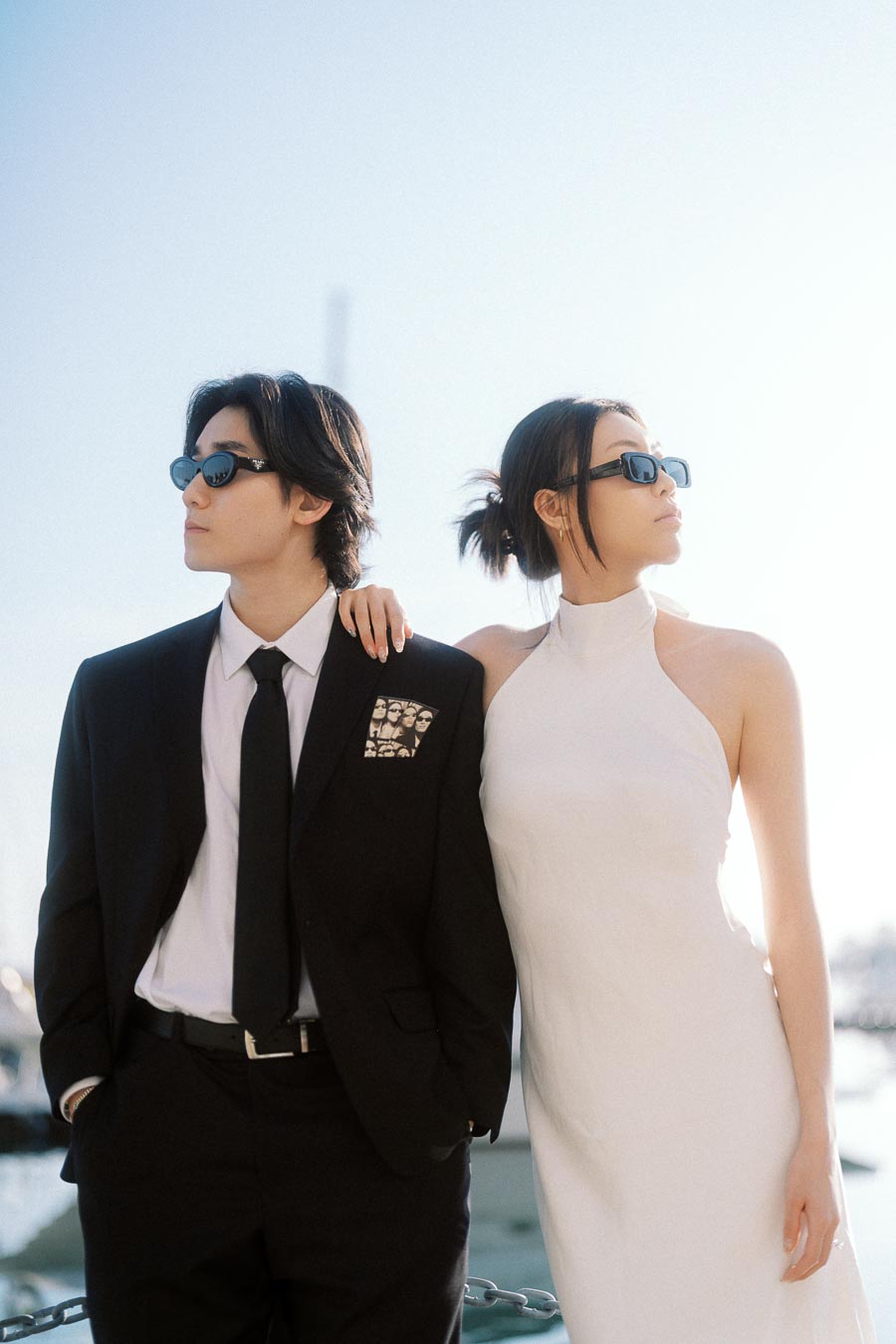 A couple wearing sunglasses, one in a black suit and the other in a white dress, standing close together by a waterfront marina in soft sunlight.
