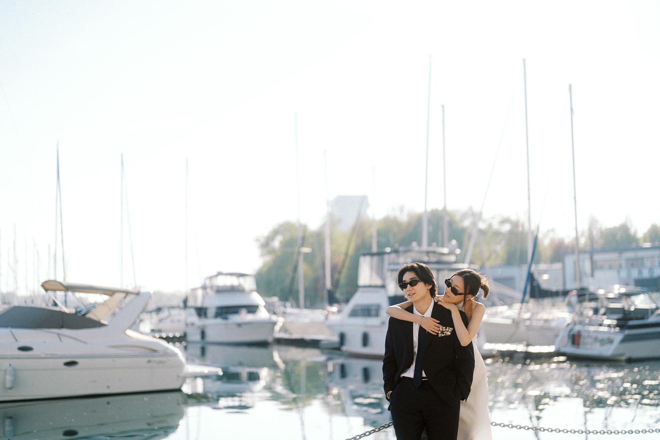 Couple in formal attire with sunglasses embracing by a marina, boats and water in the background, soft sunset light.