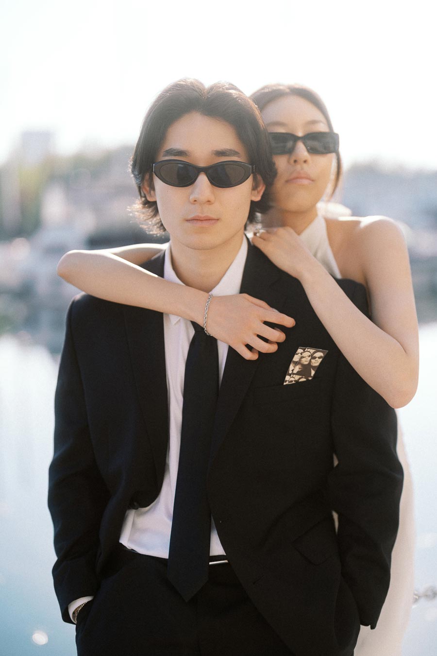 Couple wearing sunglasses embracing in modern black and white outfits, standing closely together by a sunlit waterfront setting.