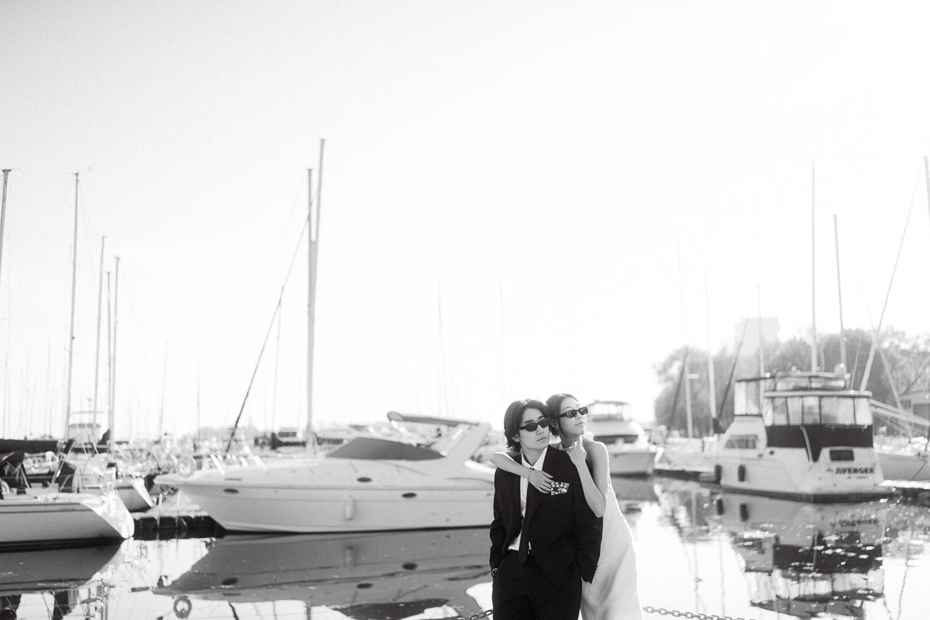 Couple in formal attire and sunglasses embracing near moored boats at a marina in soft sunset light.