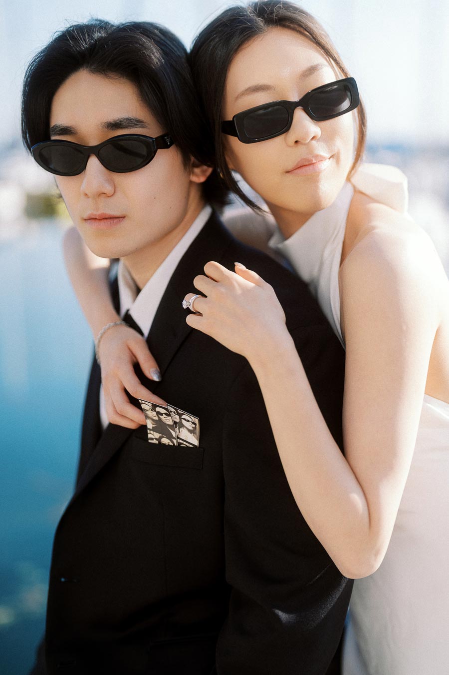 Couple wearing black sunglasses embracing, woman in white dress and man in black suit, waterfront background visible.