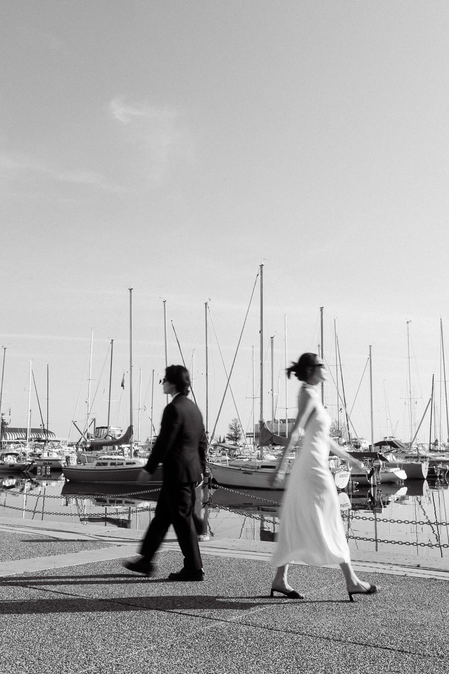 Two people in formal outfits walking along a marina with sailboats, captured in black and white on a sunny day.