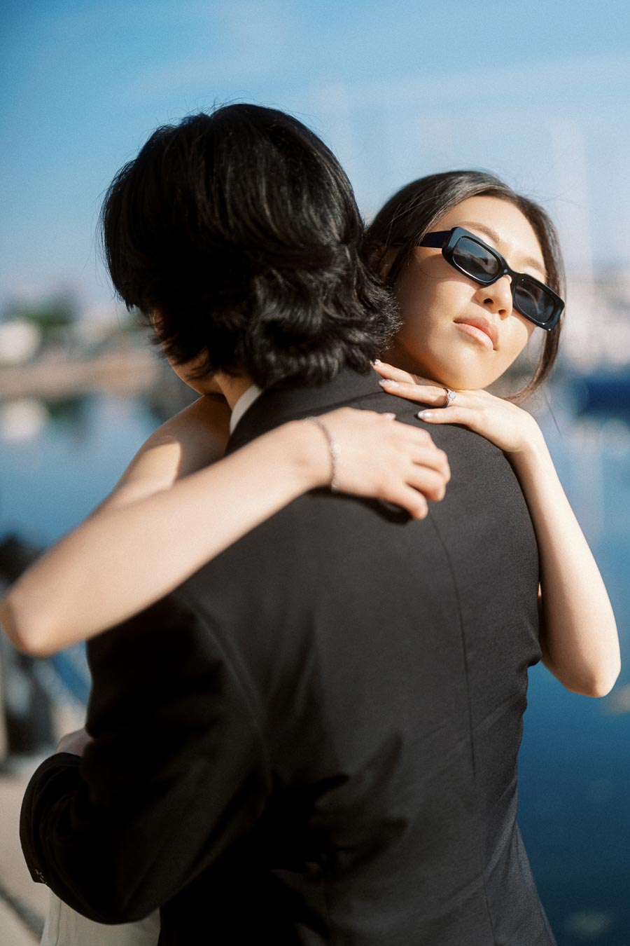 Couple embracing closely by a waterfront marina, woman in sunglasses and white dress, sunlight reflecting on water.