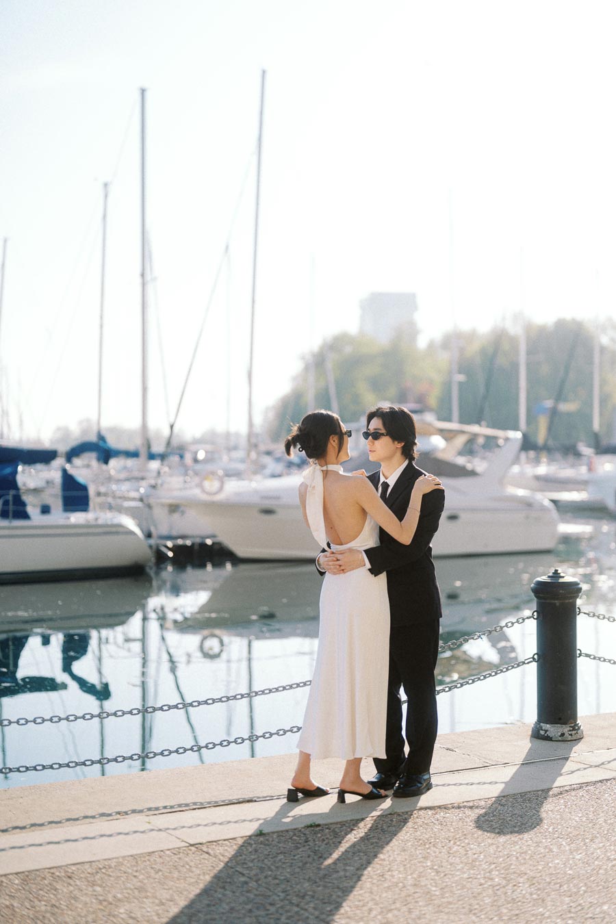 A stylish couple in formal outfits embrace by the marina, surrounded by sailboats and soft evening sunlight reflecting on the water.